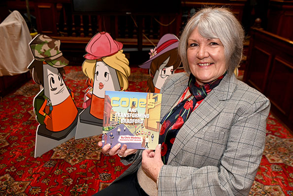 The image shows the author holding a book with some of the Cones characters in the background.