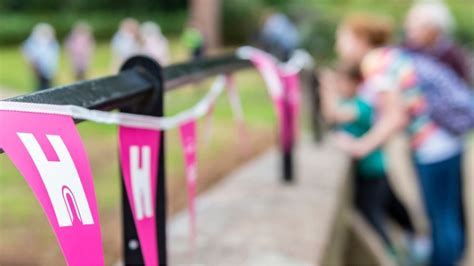 The image shows pink bunting flags with the letter H in white used by Heritage Open Days Festival. In the background is a blurred image of a family watching an activity taking place in a field.