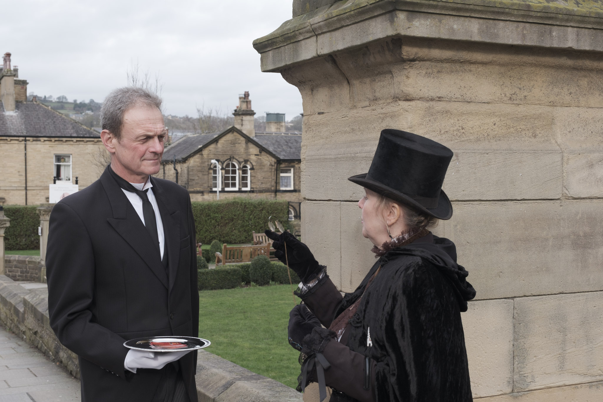 Two people in Victorian-era costume stand outside a historic stone building. A man dressed as a butler, wearing a black tailcoat, white gloves, and carrying a silver tray, listens attentively to a woman in a black velvet cape and tall top hat. The woman gestures while holding a pair of spectacles. Behind them are neatly kept gardens, stone buildings, and benches, creating the atmosphere of a historical reenactment or heritage event.