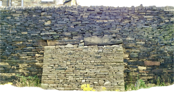 Photograph of a traditional dry stone wall constructed from stacked flat stones without mortar. In the centre of the wall is a rectangular section of finer stonework, suggesting a repaired area or infilled opening. Wildflowers and grasses grow at the base of the wall, while the roofs and upper walls of nearby stone buildings are visible behind it. The image highlights the craftsmanship and texture of traditional dry stone walling in a rural setting.