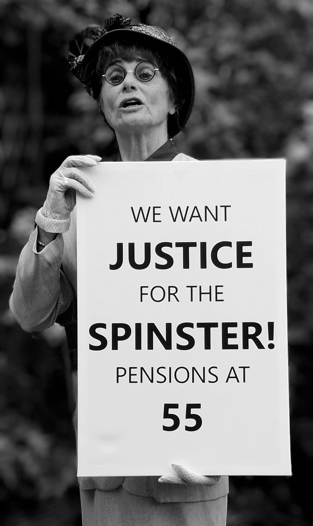 Black-and-white photograph of a woman dressed in vintage-style clothing, including a hat, round glasses, and gloves, holding a protest sign. The sign reads: “WE WANT JUSTICE FOR THE SPINSTER! PENSIONS AT 55.” She appears to be speaking or addressing a crowd, with a blurred outdoor background of trees behind her. The image evokes a historical or theatrical campaign for women's rights and social equality.