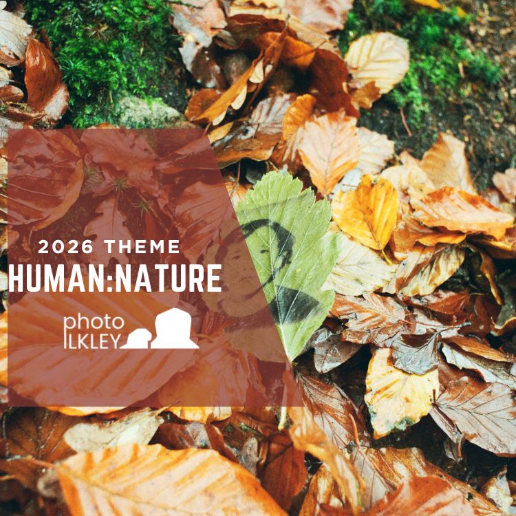 Promotional graphic for Photo Ilkley’s 2026 theme, “Human:Nature.” The image shows a woodland floor covered with fallen autumn leaves in shades of orange, brown, and gold, with patches of green moss visible between them. A green leaf near the centre features a faint human silhouette or shadow, reflecting the theme’s exploration of the relationship between people and the natural world. A translucent brown overlay contains the text “2026 Theme Human:Nature” alongside the Photo Ilkley logo.