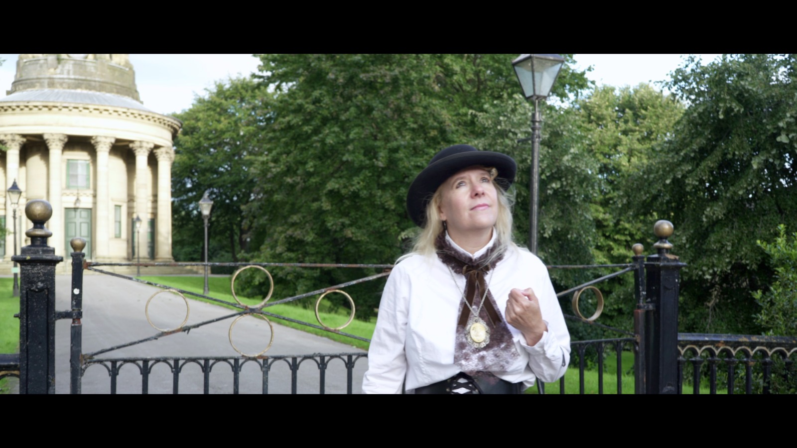 A person in historical-style costume stands in front of an ornate wrought-iron gate, looking thoughtfully upward. They wear a black hat, a white blouse with dark velvet detailing, a corset-style belt, and a pendant necklace. Behind them is a circular neoclassical building with columns, surrounded by lush green trees and parkland. The scene is outdoors on a bright day and has a theatrical, period-drama feel.