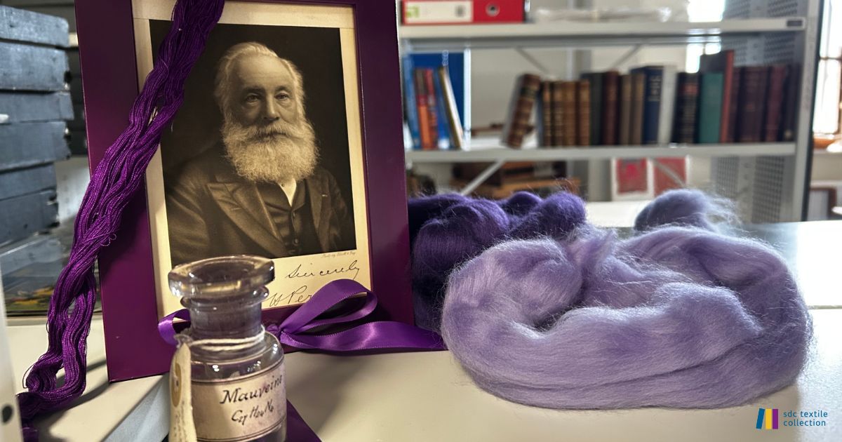 Photograph of a museum or archive display about mauve dye. In the foreground, a framed portrait of a bearded Victorian man stands beside a small glass bottle labelled “Mauveine,” with strands of deep purple yarn draped around the frame. Soft bundles of wool roving in shades of lilac and purple are arranged on a table. Bookshelves and archival storage materials are visible in the background. The display highlights the history of the synthetic purple dye mauveine and its impact on the textile industry.