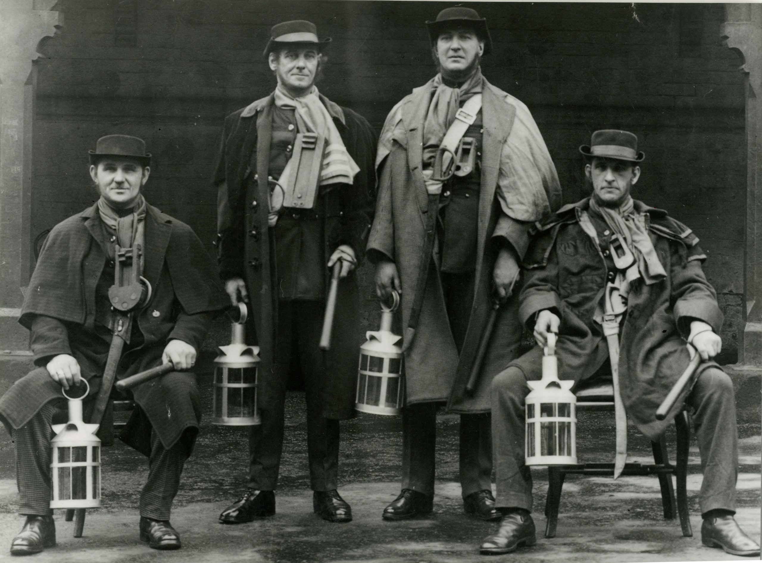 Black and white historical photograph of four Bradford night watchmen wearing long coats and hats, holding lanterns and rattles while posed outside a building.