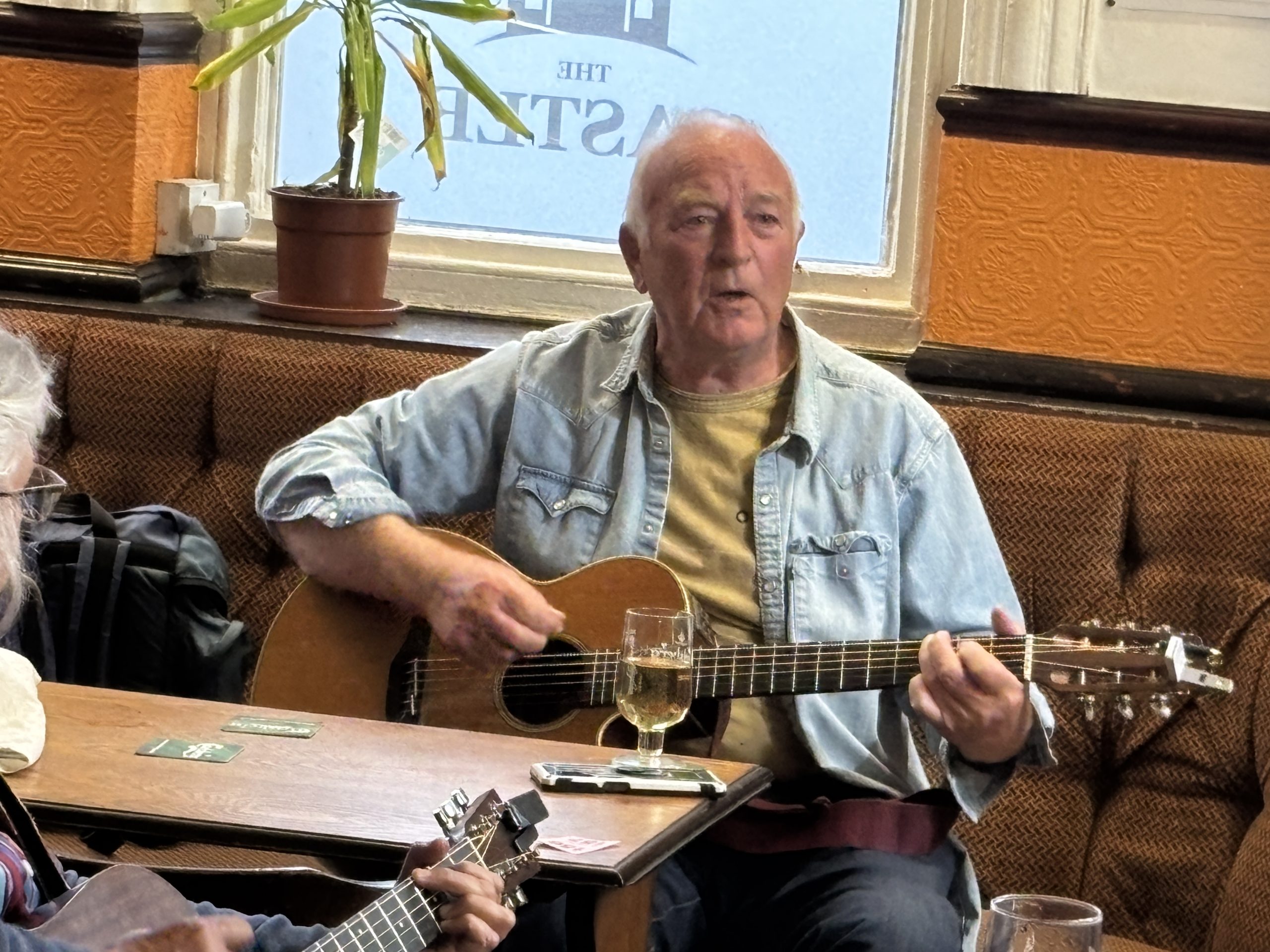Photograph of an older man seated in a pub, singing while playing an acoustic guitar. He wears a light denim shirt over a T-shirt and sits at a wooden table with a glass of drink in front of him. Other musicians holding stringed instruments are partially visible around him, suggesting an informal folk music session. The pub interior features patterned seating, orange walls, a potted plant on the windowsill, and a frosted window in the background. The image captures a relaxed community music gathering.