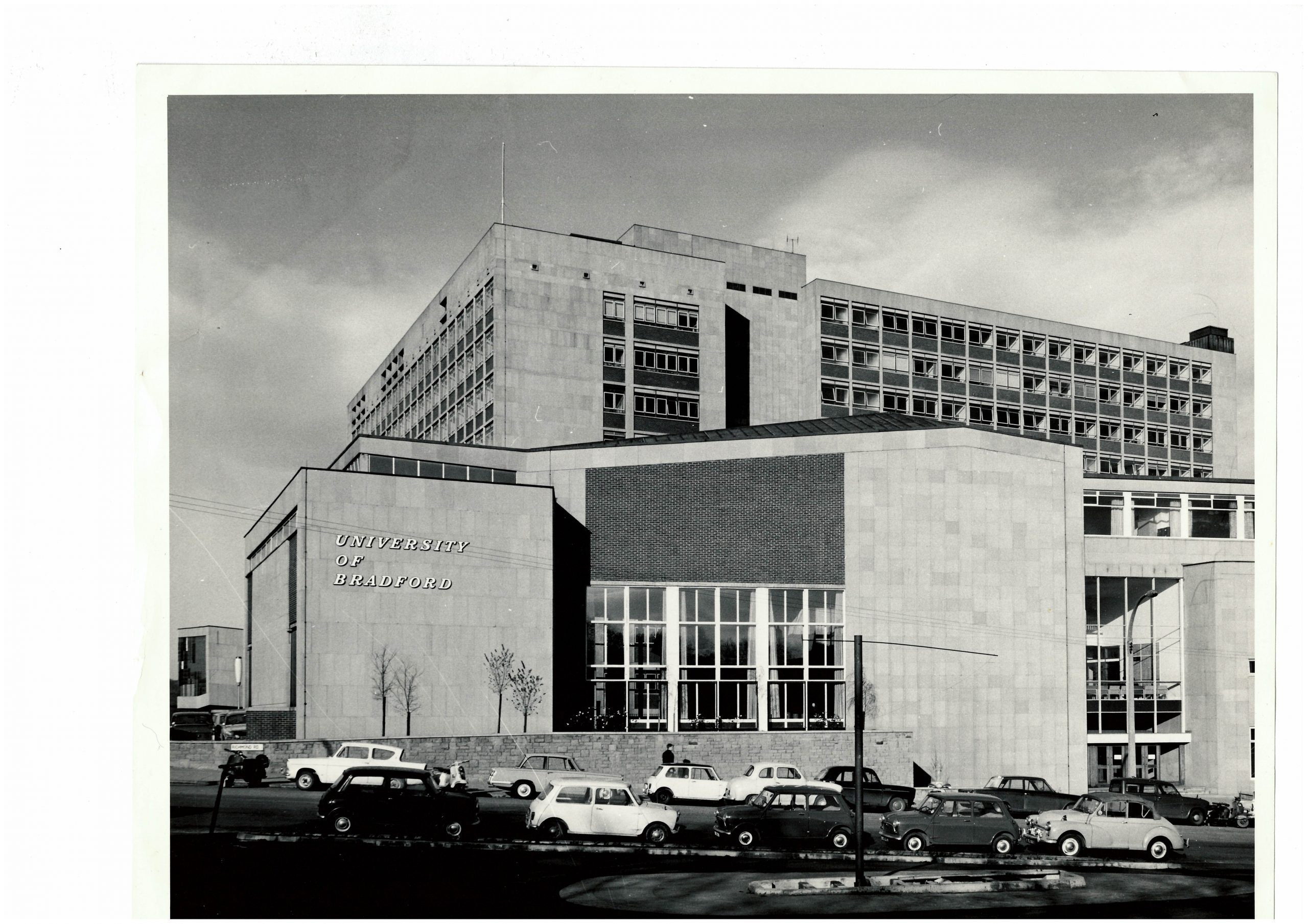 Black-and-white photograph of the University of Bradford campus in 1967. A large modernist concrete university building dominates the image, with long rows of windows and the words “University of Bradford” displayed on the front façade. Several vintage cars from the 1960s are parked along the road in the foreground, while cloudy skies frame the building above.