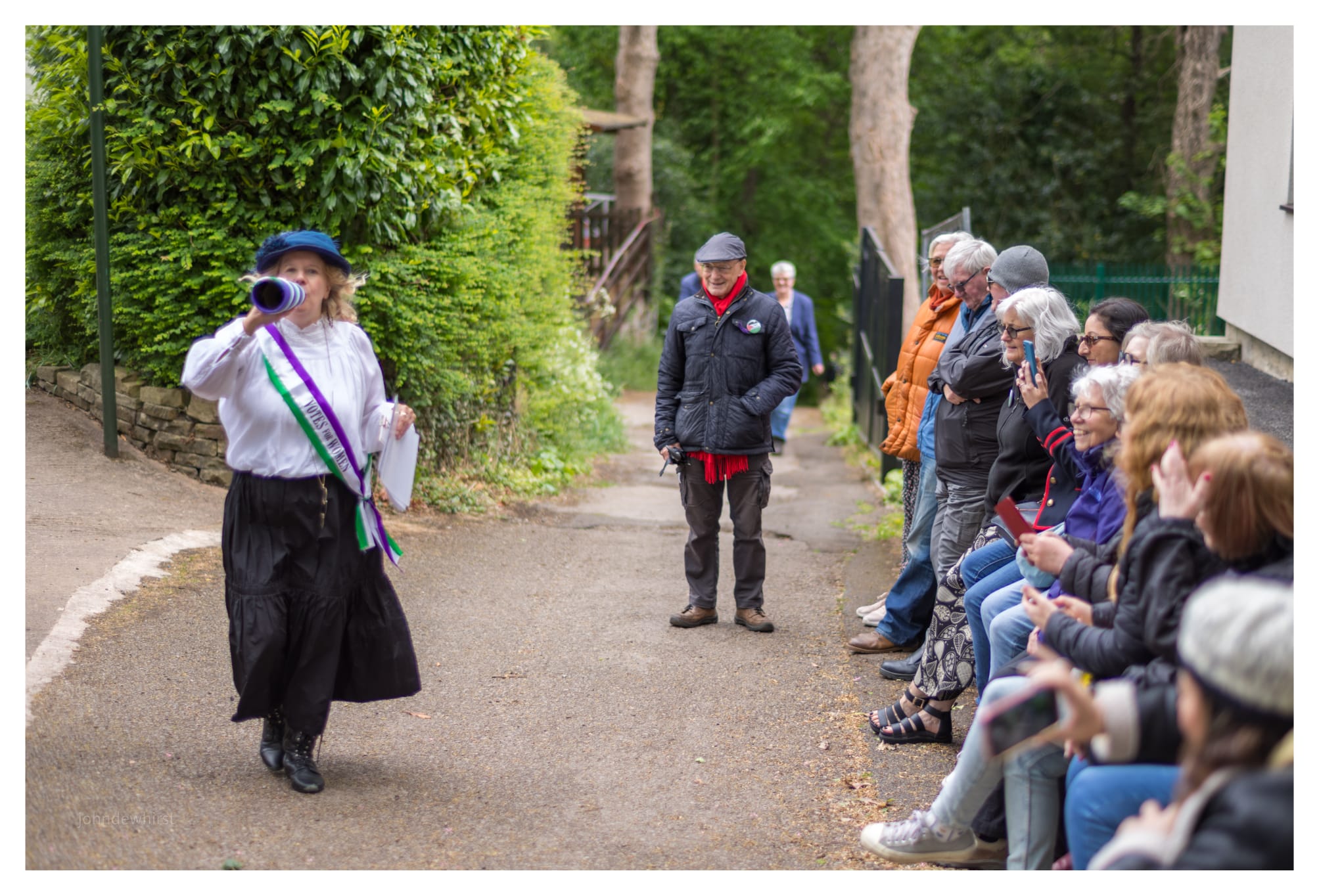 A person dressed as a suffragette walks along a tree-lined path, speaking through a blue megaphone while carrying papers. They wear a white blouse, dark skirt, blue hat, and a purple, white, and green “Votes for Women” sash. A group of spectators sit and stand along the side of the path, watching and smiling, with some taking photos on their phones. The outdoor scene is surrounded by greenery and has the atmosphere of a historical reenactment or community heritage event.