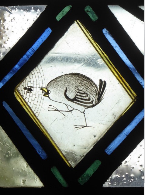 Close-up photograph of a stained-glass window panel featuring a small wren perched beside a spider’s web with a tiny spider. The bird is illustrated in fine black linework with subtle brown and yellow colouring. The central diamond-shaped panel is bordered by blue, green, and yellow geometric stained-glass pieces, with raindrops visible on the glass and a blurred outdoor landscape in the background. The image highlights the delicate craftsmanship and nature-inspired design of the stained glass.