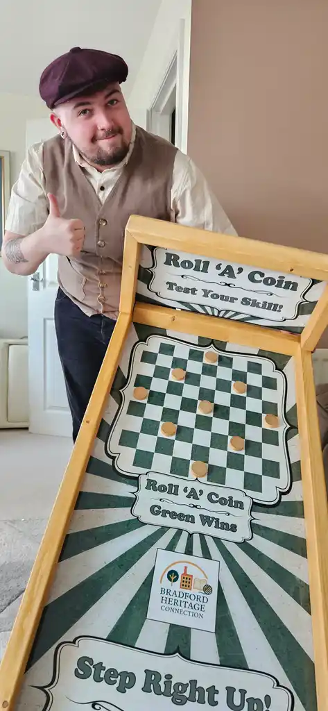A person wearing a flat cap, cream shirt, and beige waistcoat stands beside a carnival-style game board, giving a thumbs-up to the camera. The wooden game, titled “Roll A Coin – Test Your Skill,” features a green-and-white chequered target area with several coins resting on the board. Decorative vintage-style graphics and the Bradford Heritage Connection logo are displayed on the game, which is set up indoors against a neutral-coloured wall. The person smiles warmly.